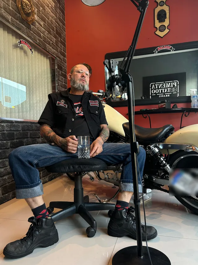 Tatuador no estúdio Pimenta Tattoo, com o letreiro da marca na parede vermelha e uma motocicleta customizada ao lado